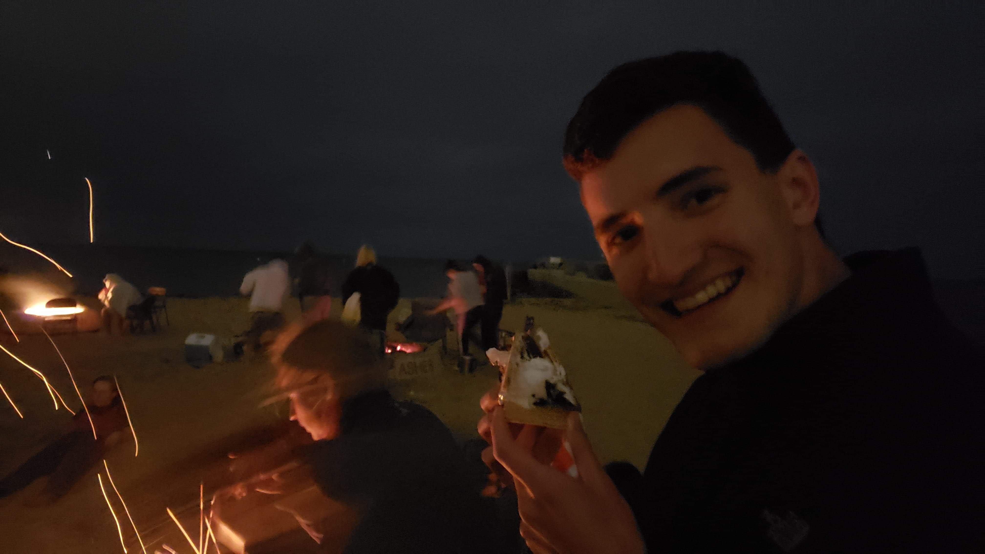 A happy Jack (before grad school) holds a s'more-like object on the beach at night.